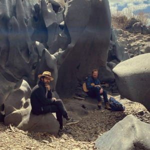 two men sitting on rocks in a canyon, one is holding a guitar and the other is striking a thinking pose